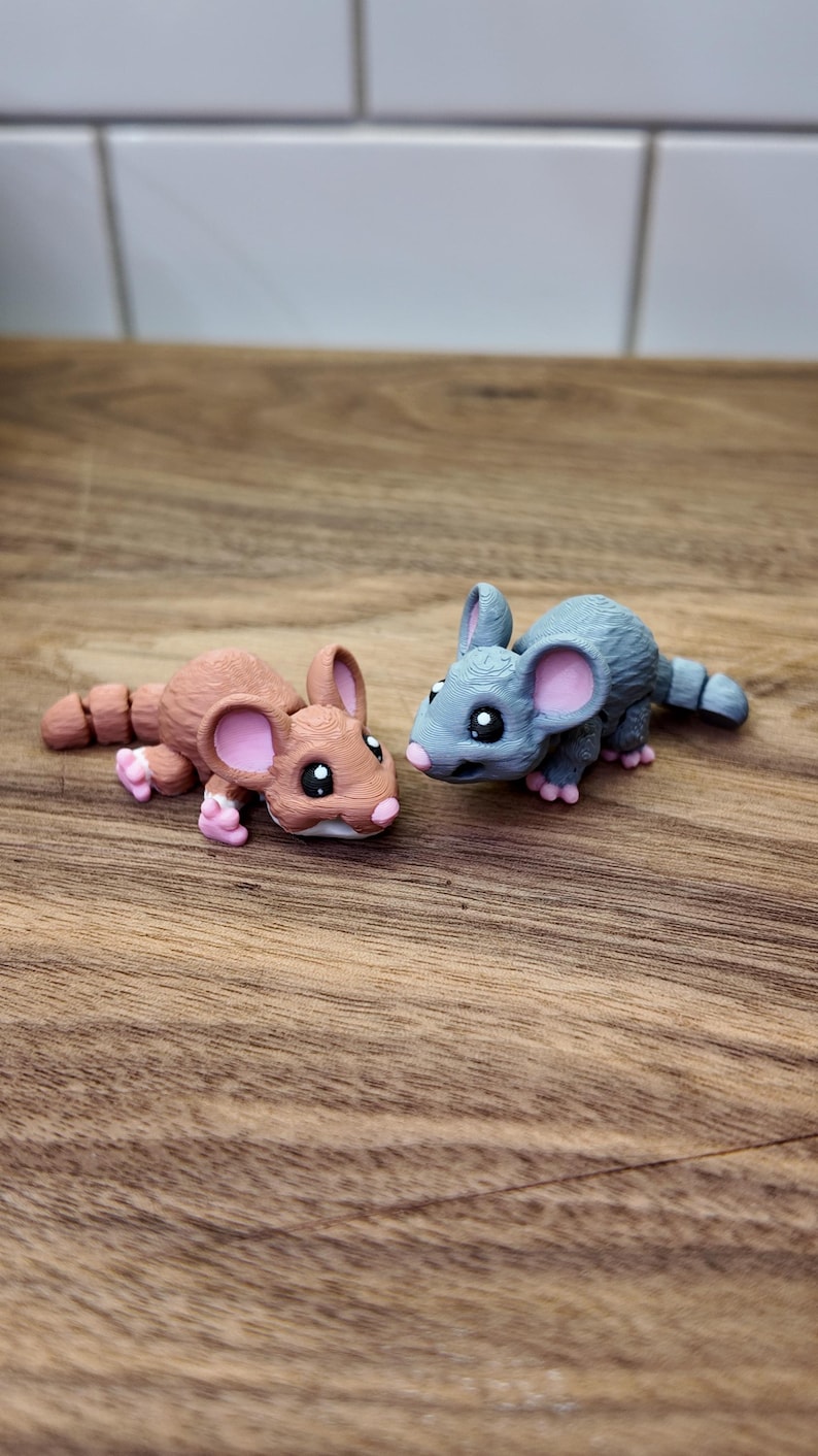 May include: Two small, detailed mouse figurines. One is brown with pink accents, the other is gray with pink accents. Both have large eyes and are lying on a wooden surface.