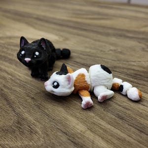 May include: Two 3D-printed cat figurines. One is black with white eyes and a pink nose, the other is white with orange and black markings. The figurines are on a wooden surface.