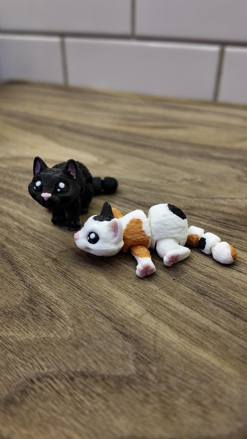 May include: Two 3D-printed cat figurines. One is black with white eyes and a pink nose, the other is white with orange and black markings. The figurines are on a wooden surface.