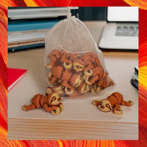 May include: A clear plastic bag filled with numerous small, brown sloth figurines. Two sloth figurines are outside the bag. The sloths have tan faces and light brown limbs. The figurines are on a white surface.