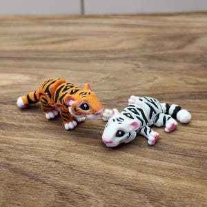 May include: Two small tiger figurines. One is orange with black stripes, the other is white with black stripes. Both have white paws and pink accents. They are posed on a wooden surface.
