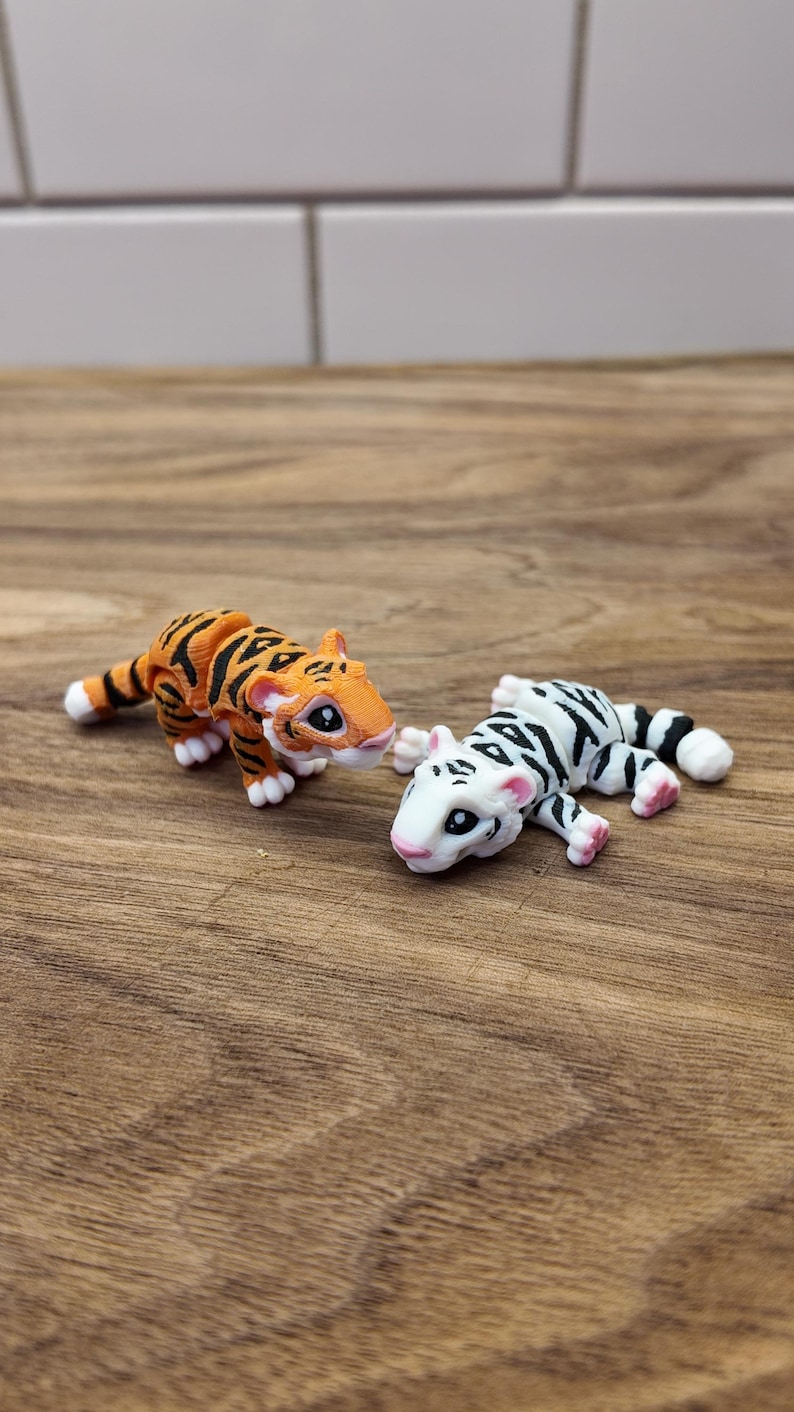 May include: Two small tiger figurines. One is orange with black stripes, the other is white with black stripes. Both have white paws and pink accents. They are posed on a wooden surface.