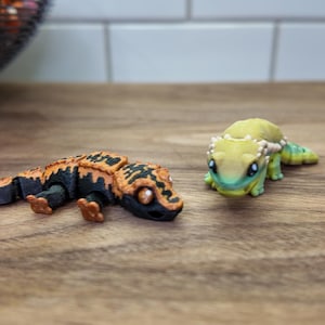 May include: Two detailed gecko figurines. One is black and orange, the other is green and yellow. The geckos are on a wooden surface. The figurines have intricate designs and are likely decorative items or toys.