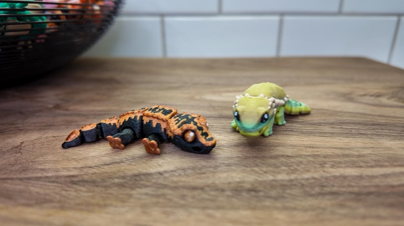 May include: Two detailed gecko figurines. One is black and orange, the other is green and yellow. The geckos are on a wooden surface. The figurines have intricate designs and are likely decorative items or toys.