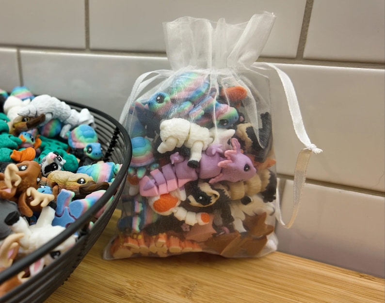 May include: A clear, drawstring bag filled with colorful, miniature animal figurines. The bag is next to a black wire basket overflowing with similar figurines. The figurines include turtles, sheep, and other creatures.