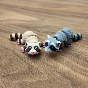 May include: Two small raccoon figurines. One is beige and brown, the other is gray and black. Both have black masks, pink paws, and striped tails. They are lying on a wooden surface.