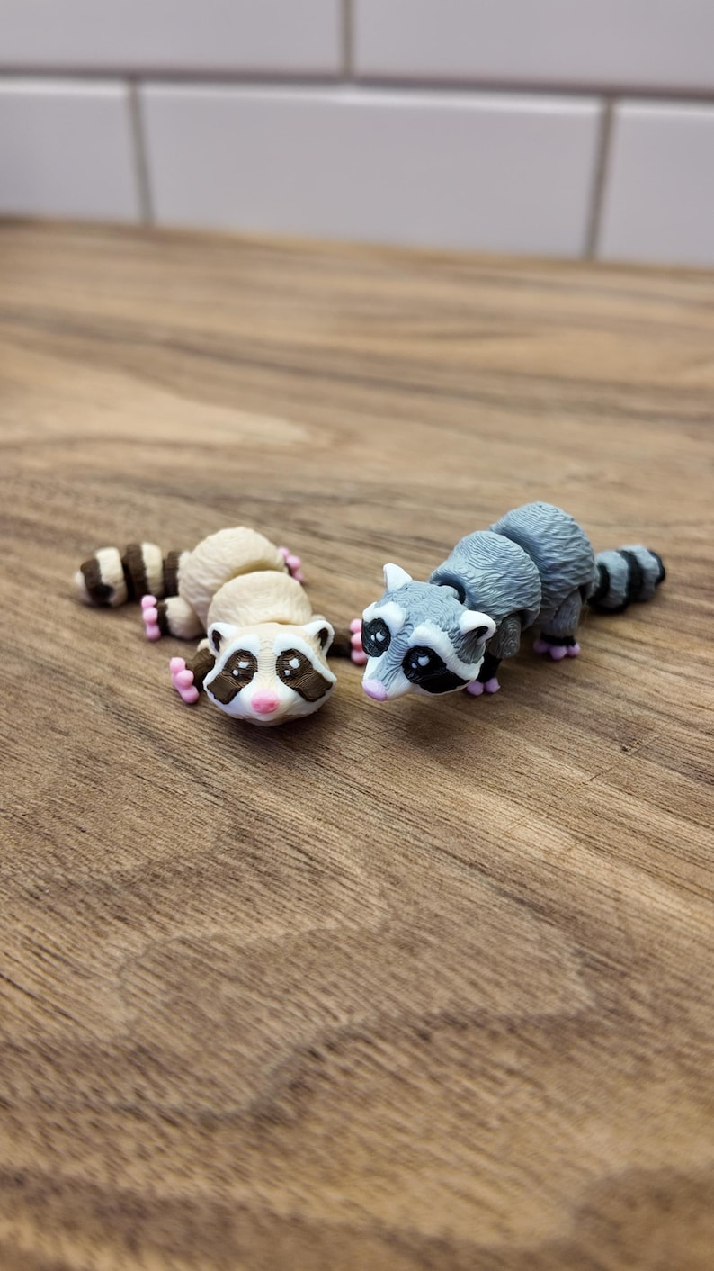 May include: Two small raccoon figurines. One is beige and brown, the other is gray and black. Both have black masks, pink paws, and striped tails. They are lying on a wooden surface.
