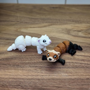 May include: Two articulated ferret figurines. One is white with pink accents, the other is brown, black, and tan. The toys are posed on a wooden surface, with a white tile background.