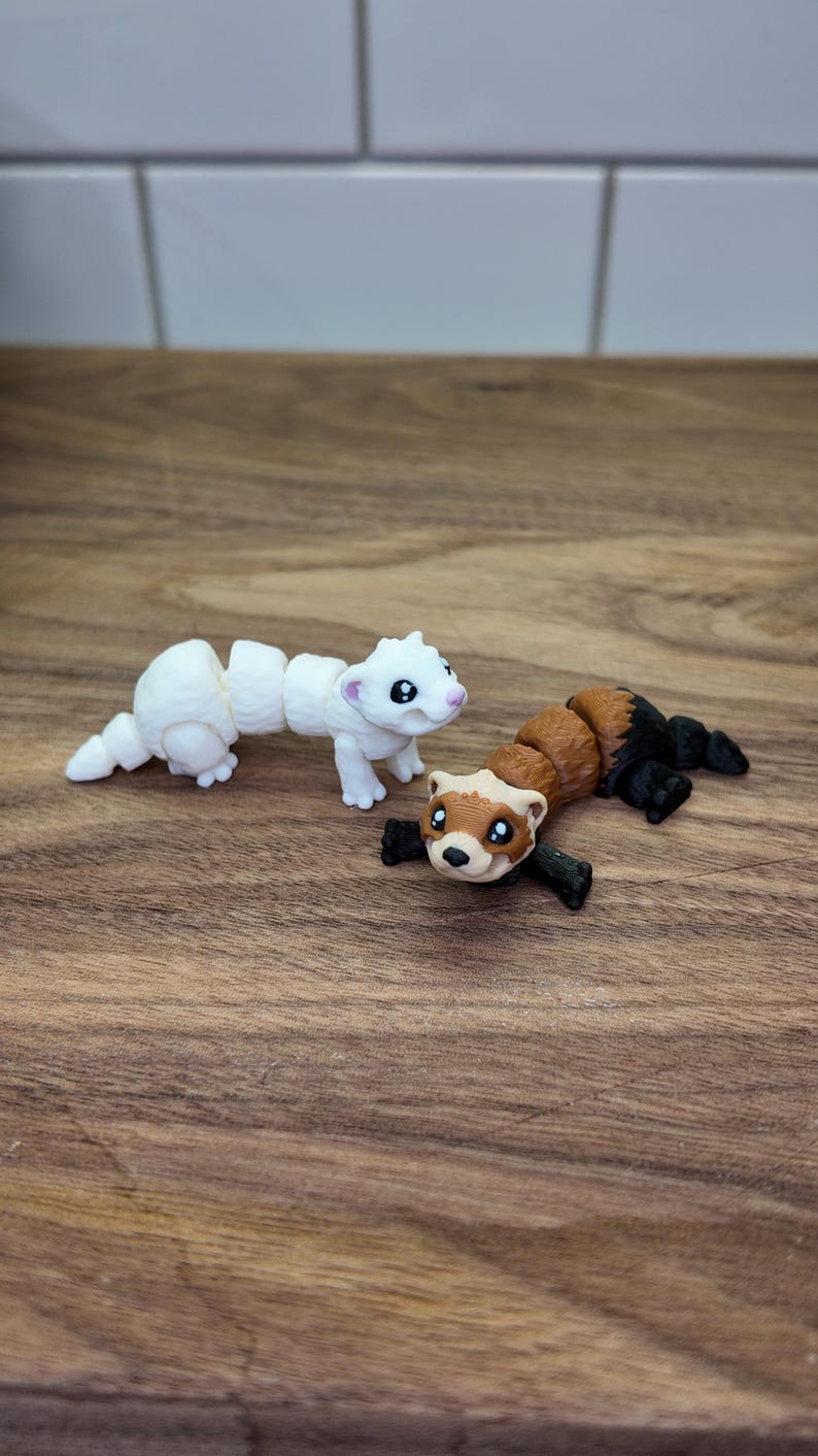 May include: Two articulated ferret figurines. One is white with pink accents, the other is brown, black, and tan. The toys are posed on a wooden surface, with a white tile background.
