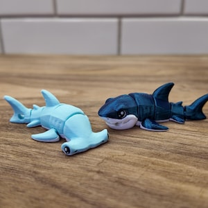May include: Two toy sharks on a wooden surface. One is a light blue hammerhead shark, and the other is a dark blue shark with white accents. The sharks are small, detailed figurines, likely for play or display.