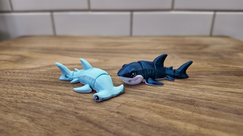 May include: Two toy sharks on a wooden surface. One is a light blue hammerhead shark, and the other is a dark blue shark with white accents. The sharks are small, detailed figurines, likely for play or display.