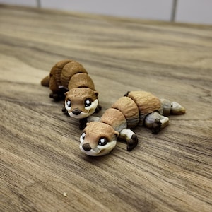 May include: Three brown and tan articulated otter figurines with black paws and white eyes are displayed on a wooden surface. The otters are segmented, allowing for movement. The background is a light-colored tiled wall.
