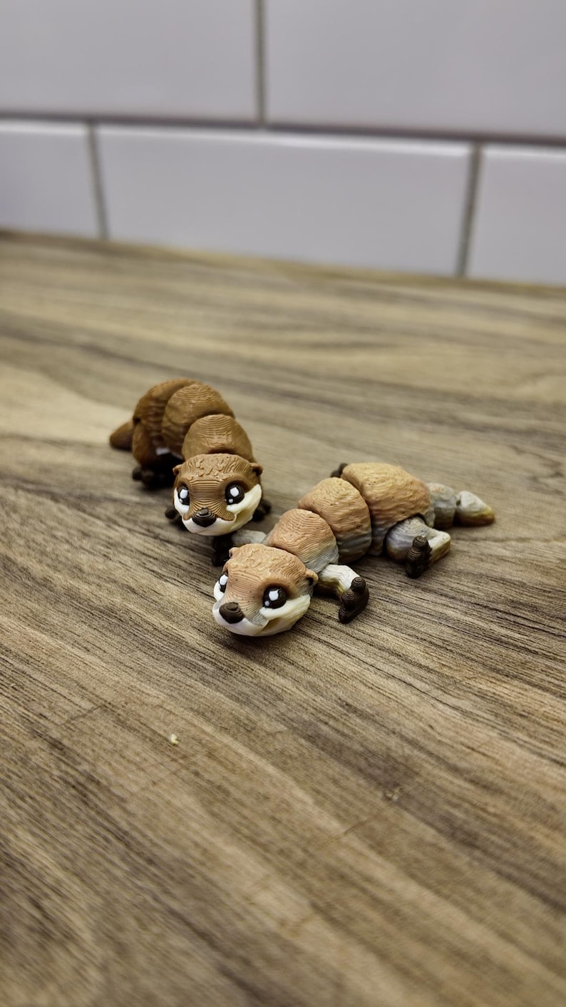 May include: Three brown and tan articulated otter figurines with black paws and white eyes are displayed on a wooden surface. The otters are segmented, allowing for movement. The background is a light-colored tiled wall.