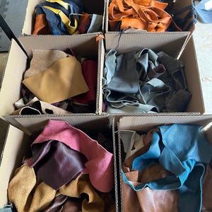 May include: Six cardboard boxes filled with assorted leather scraps in various colors including blue, orange, tan, burgundy, pink, and grey. The pieces vary in texture and size, showing smooth and suede finishes packed together in each container.