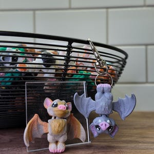 May include: Two bat-shaped figurines, one upright and one hanging upside down, with a keychain attached. The upright bat has tan and orange wings, while the hanging bat is gray. A black wire basket filled with colorful objects is in the background.