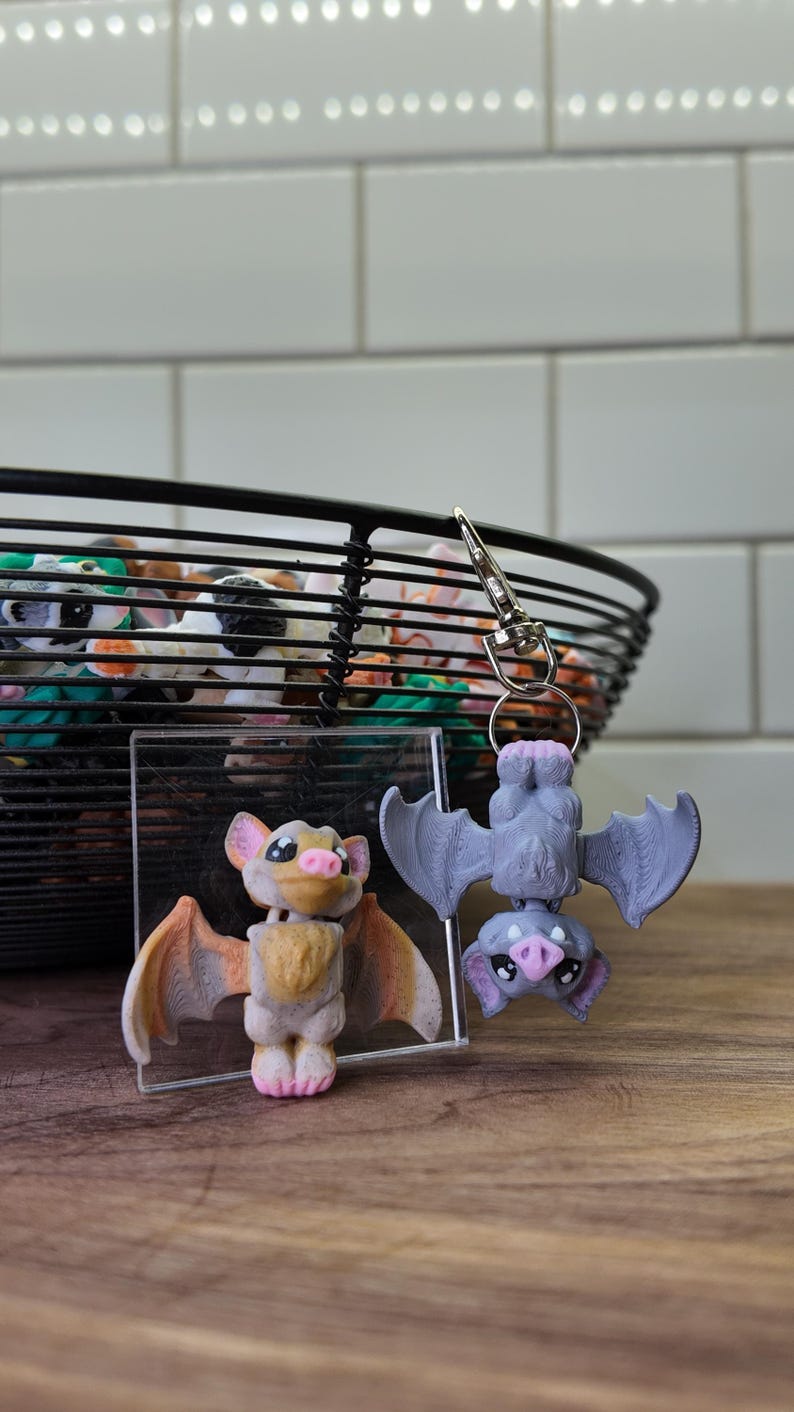 May include: Two bat-shaped figurines, one upright and one hanging upside down, with a keychain attached. The upright bat has tan and orange wings, while the hanging bat is gray. A black wire basket filled with colorful objects is in the background.