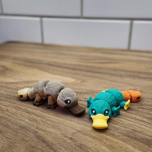 May include: Two small, detailed platypus figurines. One is tan and brown, the other is teal, orange, and yellow. They are lying on a wooden surface, with a white tile background.