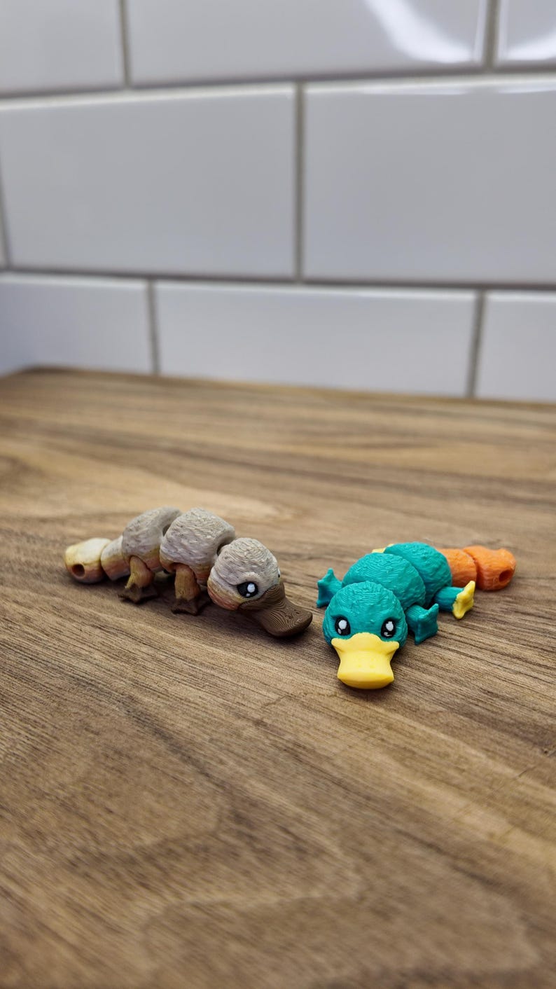 May include: Two small, detailed platypus figurines. One is tan and brown, the other is teal, orange, and yellow. They are lying on a wooden surface, with a white tile background.