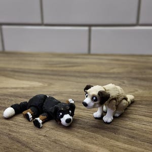 May include: Two small dog figurines. One is black and white, lying down. The other is tan and white, sitting up. Both have large, expressive eyes and are on a wooden surface.