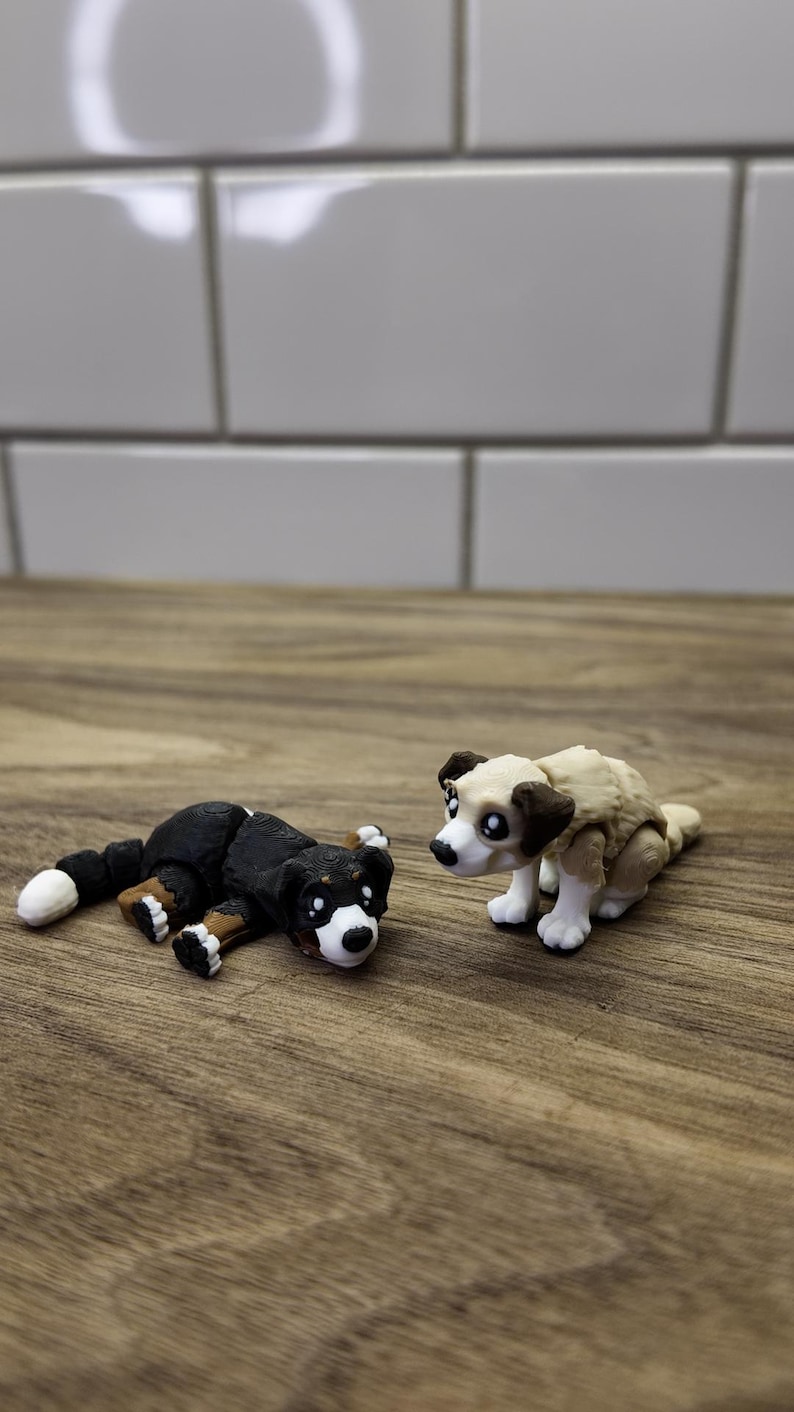 May include: Two small dog figurines. One is black and white, lying down. The other is tan and white, sitting up. Both have large, expressive eyes and are on a wooden surface.