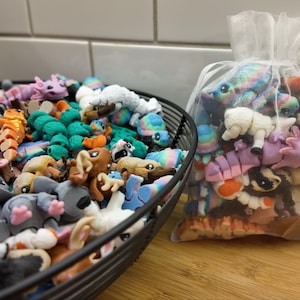 May include: A large collection of colorful, small, animal-shaped figurines in a black wire basket and a white mesh bag. The figurines are made of a soft, pliable material and are in a variety of colors, including pink, blue, green, and purple.