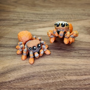 May include: Two orange and brown spider figurines with large eyes. The spiders have detailed legs and bodies, and are posed on a wooden surface. The figurines are likely handmade and are a unique decorative item.
