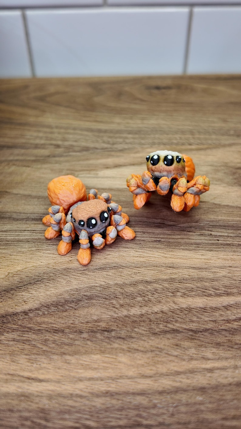 May include: Two orange and brown spider figurines with large eyes. The spiders have detailed legs and bodies, and are posed on a wooden surface. The figurines are likely handmade and are a unique decorative item.