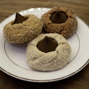 May include: Three cookies on a white plate with a silver rim. Each cookie has a chocolate candy in the center and is covered in a white, granular coating. The cookies are arranged on a wooden surface.