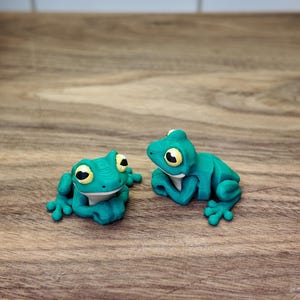 May include: Two turquoise frog figurines with yellow and black eyes rest on a wooden surface. The frogs are in a relaxed pose, with their arms and legs slightly extended. The background is a light-colored tile.