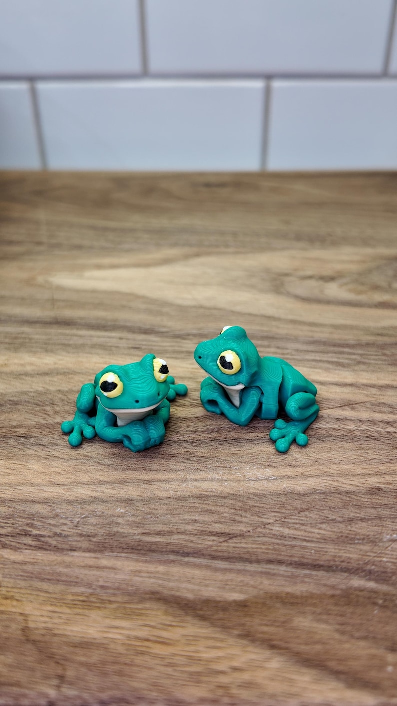 May include: Two turquoise frog figurines with yellow and black eyes rest on a wooden surface. The frogs are in a relaxed pose, with their arms and legs slightly extended. The background is a light-colored tile.