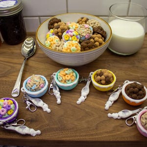 May include: A wooden cutting board displays a bowl of colorful cereal, a glass of milk, and several miniature cereal bowl keychains. Jars of jam and a spoon are also on the board. The keychains feature various cereal types in small bowls.
