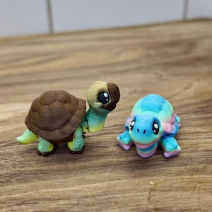 May include: Two small, colorful turtle figurines. One has a brown shell and a head with brown and tan coloring. The other turtle has a shell with blue, pink, and green hues. Both have detailed features and are set on a wooden surface.