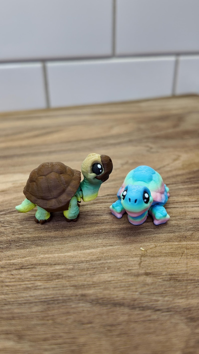 May include: Two small, colorful turtle figurines. One has a brown shell and a head with brown and tan coloring. The other turtle has a shell with blue, pink, and green hues. Both have detailed features and are set on a wooden surface.
