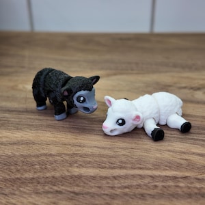 May include: Two small animal figurines: a black lamb and a white lamb. The black lamb stands upright, while the white lamb lies down. Both have large eyes and are set on a wooden surface.