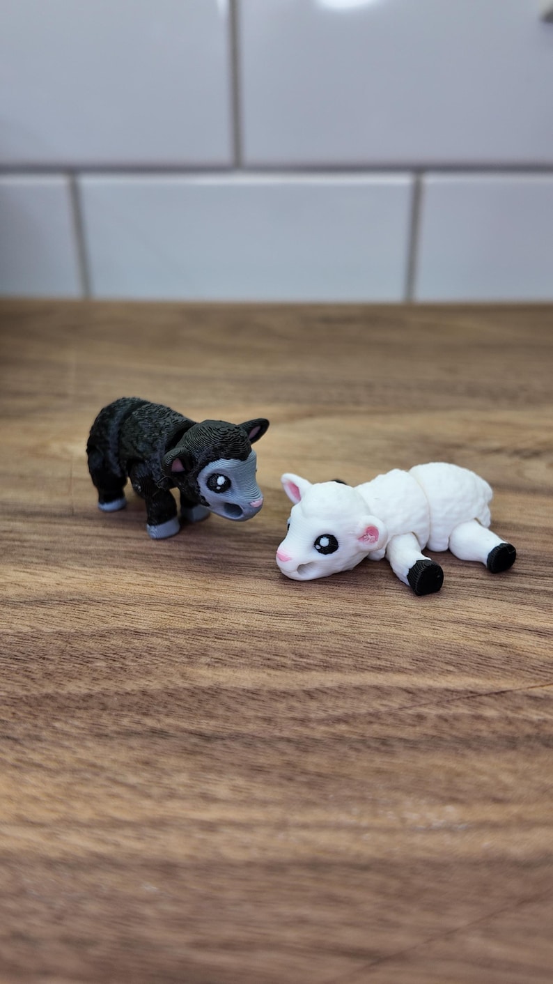 May include: Two small animal figurines: a black lamb and a white lamb. The black lamb stands upright, while the white lamb lies down. Both have large eyes and are set on a wooden surface.