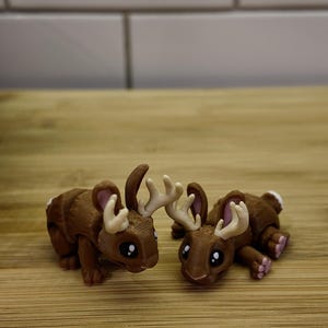 May include: Two brown figurines, each a hybrid of a rabbit and a moose, with antlers and bunny ears. The creatures have large black eyes and pink accents on their paws and ears. They are lying down on a wooden surface.