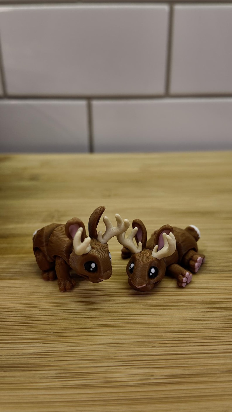 May include: Two brown figurines, each a hybrid of a rabbit and a moose, with antlers and bunny ears. The creatures have large black eyes and pink accents on their paws and ears. They are lying down on a wooden surface.
