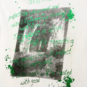 May include: White t-shirt with a green and black graphic print. The graphic features a blurred image of a waterfall and handwritten text in green. The text includes phrases like "protect the environment" and "people's hands."