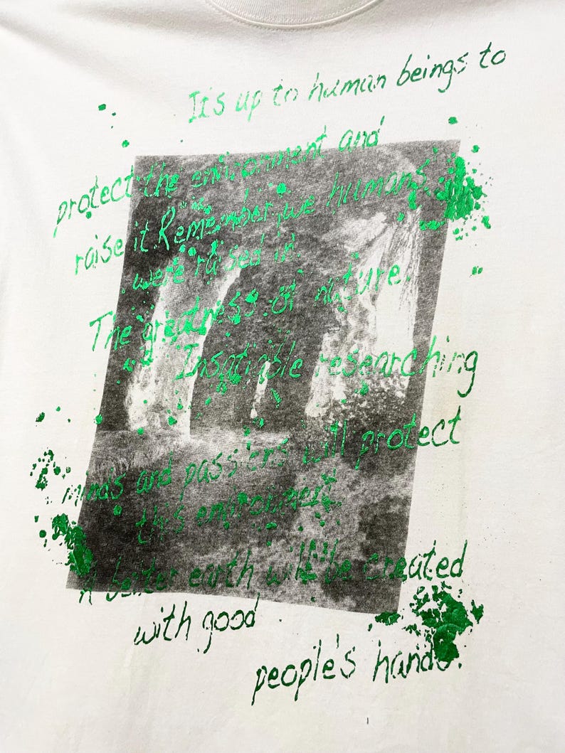 May include: White t-shirt with a green and black graphic print. The graphic features a blurred image of a waterfall and handwritten text in green. The text includes phrases like "protect the environment" and "people's hands."