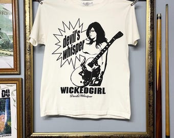 Può includere: T-shirt color crema con grafica nera di una donna che suona la chitarra, con la scritta "devil's whisper" in una stella e "WICKEDGIRL" sotto. La maglietta è appesa a un filo all'interno di una cornice dorata.