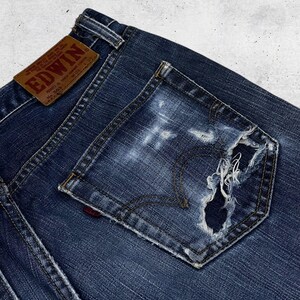 May include: Close-up of a pair of dark blue denim jeans with a distressed pocket. The jeans feature a brown leather patch with the brand name "EDWIN" and a red logo on the pocket. The denim has a worn, faded appearance.