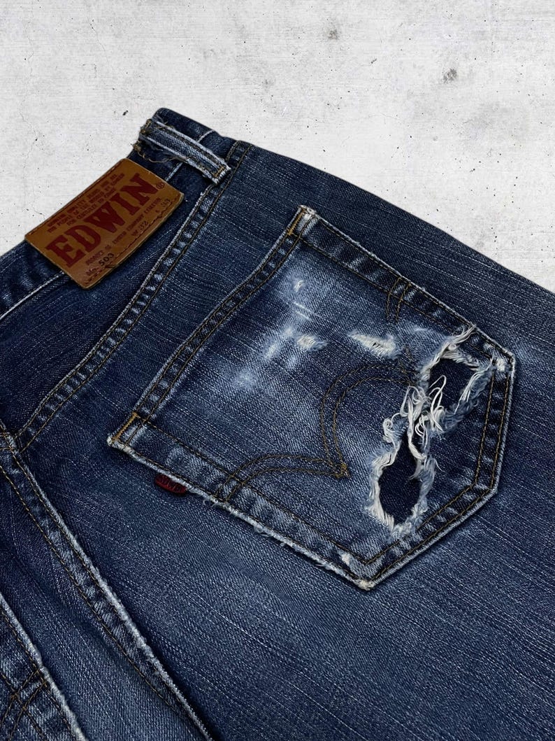 May include: Close-up of a pair of dark blue denim jeans with a distressed pocket. The jeans feature a brown leather patch with the brand name "EDWIN" and a red logo on the pocket. The denim has a worn, faded appearance.