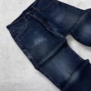 May include: Dark blue denim jeans with a classic five-pocket design. The jeans have a faded appearance and a button and zipper closure. The denim is a medium weight, suitable for everyday wear. The jeans are laid flat on a light gray surface.
