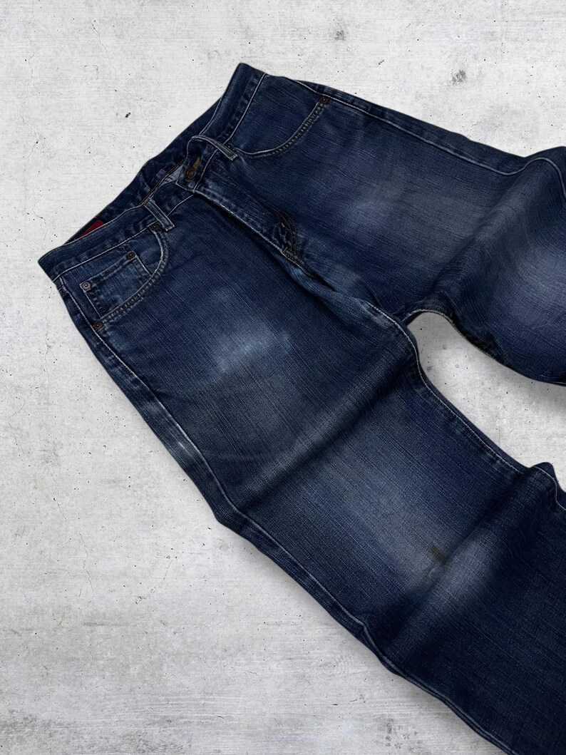 May include: Dark blue denim jeans with a classic five-pocket design. The jeans have a faded appearance and a button and zipper closure. The denim is a medium weight, suitable for everyday wear. The jeans are laid flat on a light gray surface.