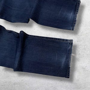 May include: Dark blue denim jeans, folded and laid out. The jeans have a classic five-pocket design and a straight leg cut. The denim has a slightly faded appearance, with visible wear and tear on the edges.