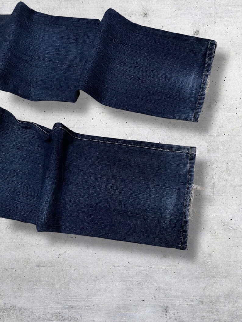 May include: Dark blue denim jeans, folded and laid out. The jeans have a classic five-pocket design and a straight leg cut. The denim has a slightly faded appearance, with visible wear and tear on the edges.