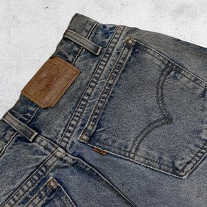 May include: Close-up of light blue denim jeans. The jeans feature a brown leather patch with text, a back pocket, and visible stitching. The jeans are a vintage style.