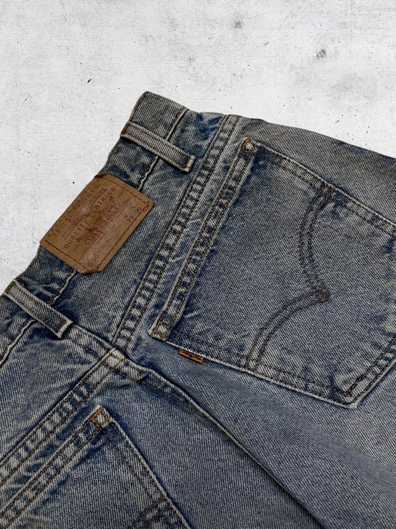 May include: Close-up of light blue denim jeans. The jeans feature a brown leather patch with text, a back pocket, and visible stitching. The jeans are a vintage style.