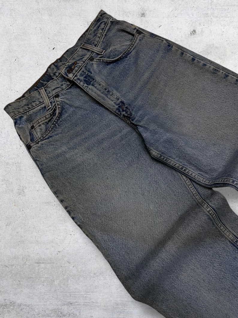 May include: Faded blue denim jeans with classic five-pocket styling. The jeans have a button and zipper closure, belt loops, and a worn appearance. The denim is a medium wash with subtle fading and wear marks.