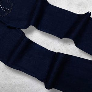 Puede incluir: Vaqueros de mezclilla azul oscuro con un dise&ntilde;o cl&aacute;sico de cinco bolsillos. El bolsillo trasero presenta un patr&oacute;n decorativo de peque&ntilde;os tachuelas plateadas. Los vaqueros est&aacute;n doblados y colocados planos, mostrando la textura y el color de la tela.